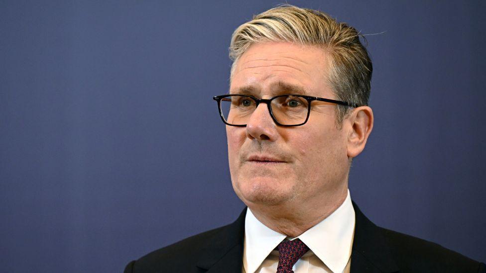 Keir Starmer, who has grey hair and glasses, wearing a dark suit, white shirt and purple tie. He is standing in front of a blue wall.