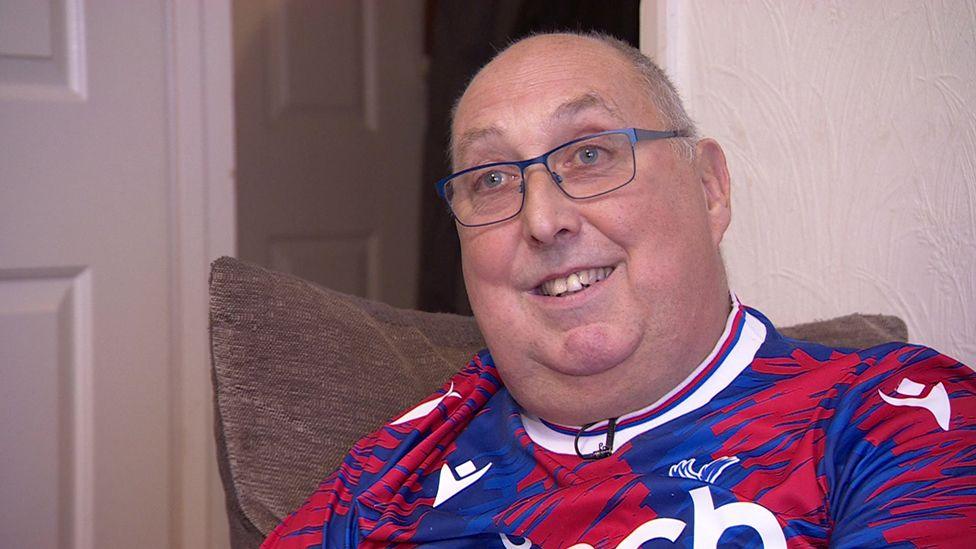 Andrew Frost, with a receding hairline and wearing glasses, is sat at home on a chair and is wearing a blue and red football shirt