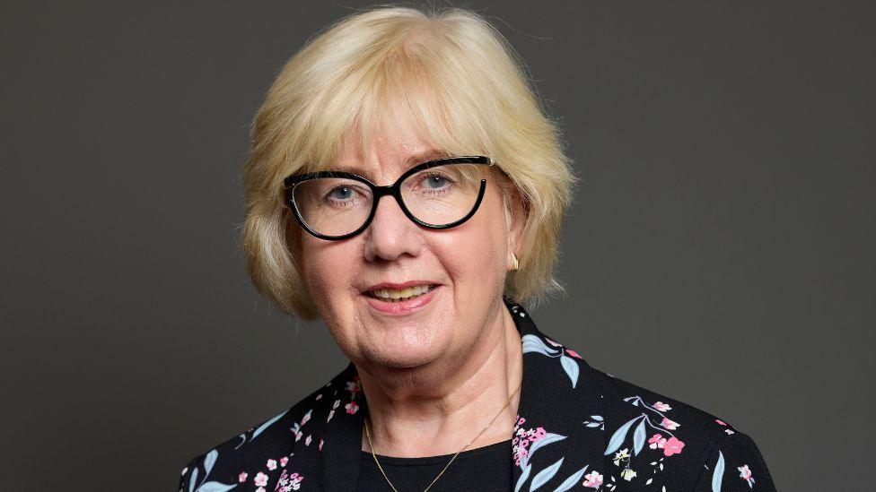 A woman with blonde hair and glasses in a close-up shot. She is looking at the camera and is visible from the shoulders up. She is wearing a dark jacket with a floral pattern. She is in front of a grey background.