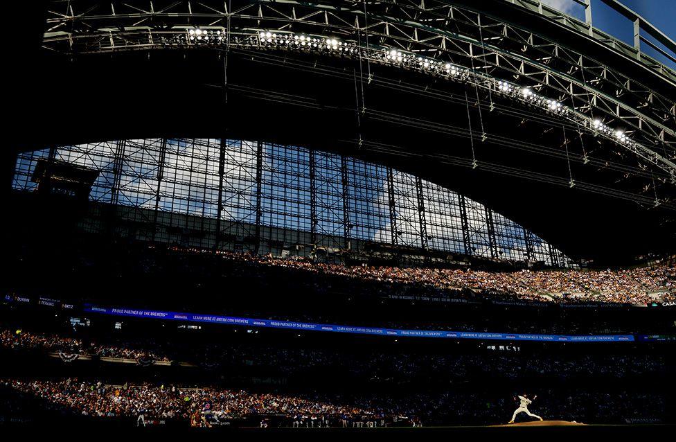 Milwaukee Brewers’ Aaron Ashby delivers a pitch against the Chicago Cubs during Game One of the National League Division Series in Wisconsin. The image shows Ashby on a sunlit mound inside a large stadium with a partially open retractable roof, contrasting bright sky and shadowed stands filled with spectators.