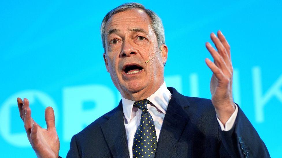 Nigel Farage, a tanned man with grey hair, speaks with his arms raised in front of a blue background. He is wearing a dark suit, white shirt and spotty tie. 