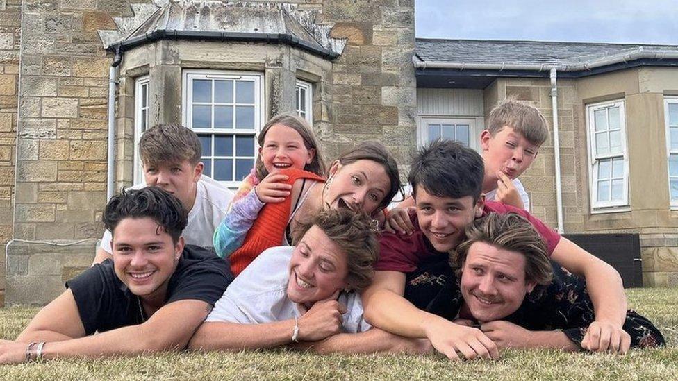 Eight young people lie on the grass piled on top of each other. Six boys and two girls, children and teenagers smiling and pulling faces for the camera. Windows of old sandstone house behind them.