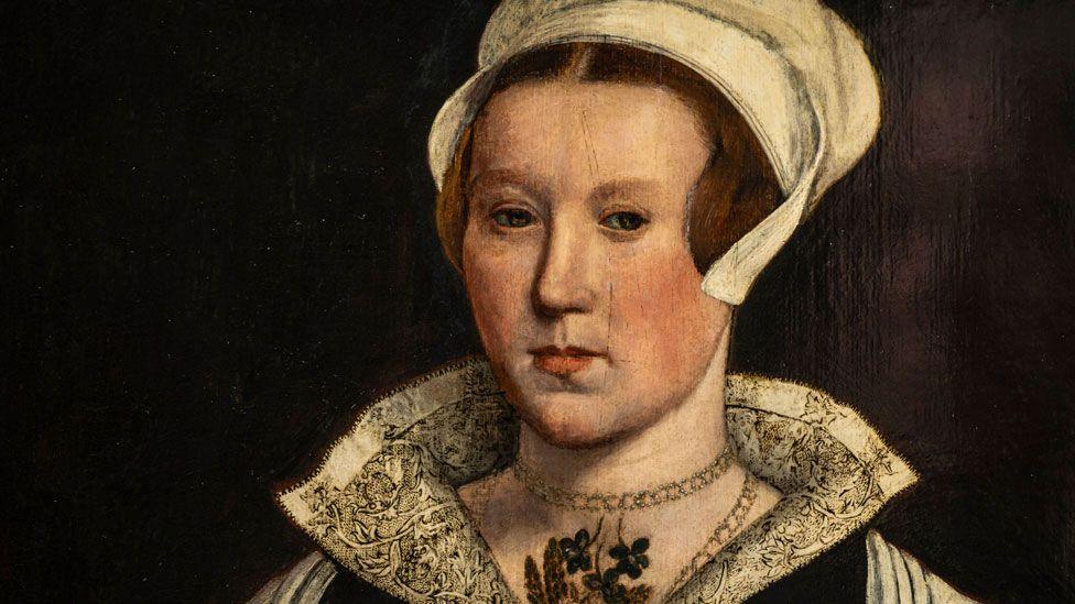 A portrait of a Tudor woman believed to be Lady Jane Grey. She has reddish hair under a white linen cap, a white lacework open collar,  raised about her neck and a gold necklace looped around her neck. She is looking left.