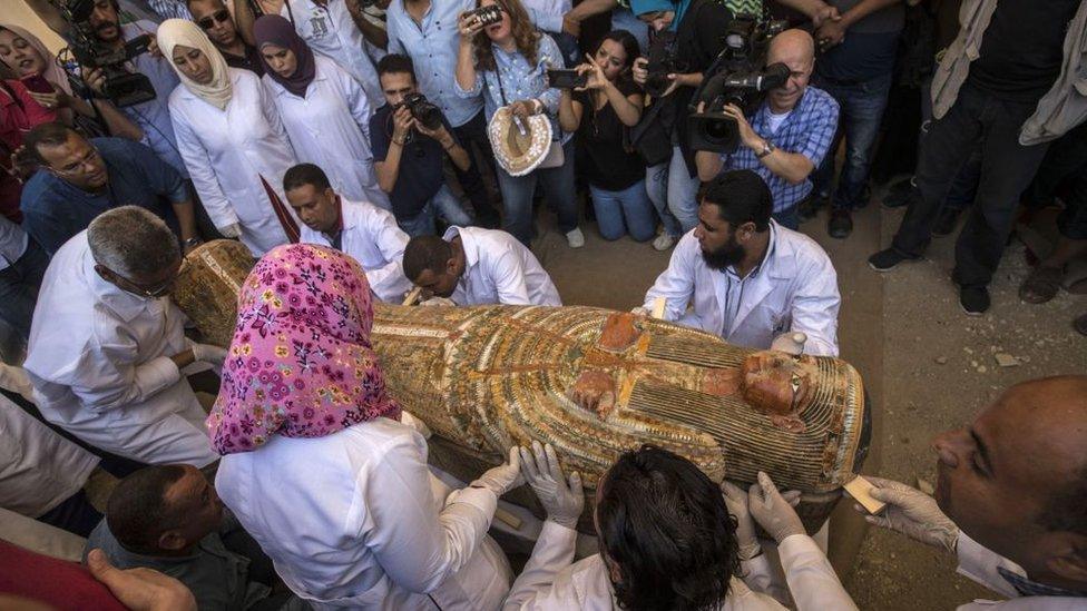 Expert-closely-studies-a-sarcophagus-as-media-crowd-around