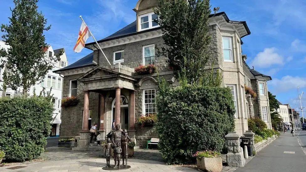 The exterior of the States Offices on a blue sky day. There are sculptures of a man, woman and two children in the square in front of the building. There is a tree, bush and granite planter of flowers either side of the entrance. The entrance has a portico with columns which has the Guernsey flag flying from it. The building has three storeys and stands next to a road.