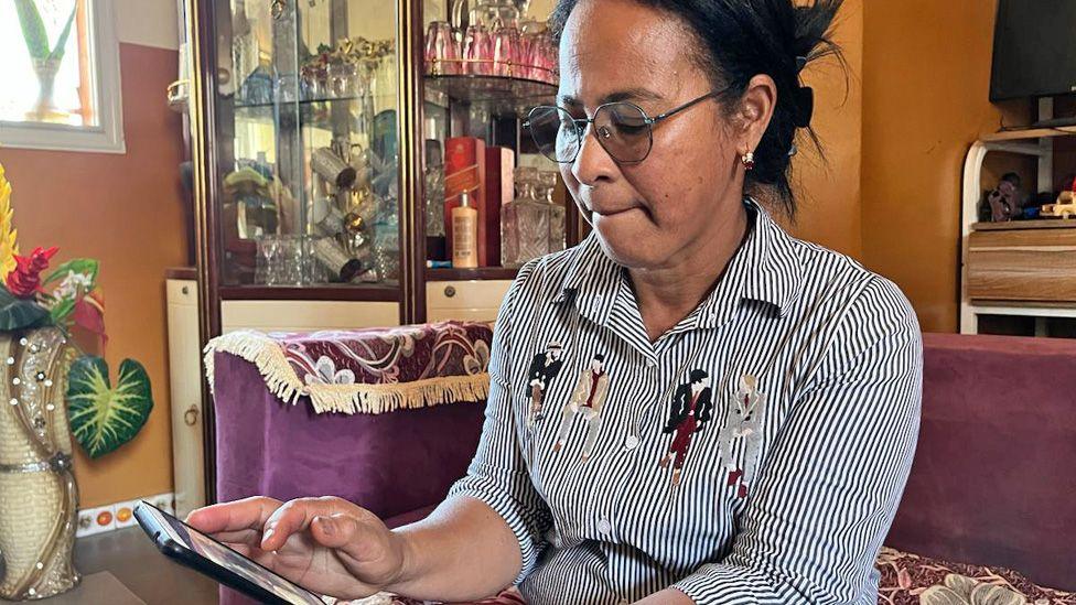 Razanasoa Edmondine, sucking her lips, in a black and white stripy long-sleeved shirt looking at the last photos of her grandson at her home near Antananarivo.