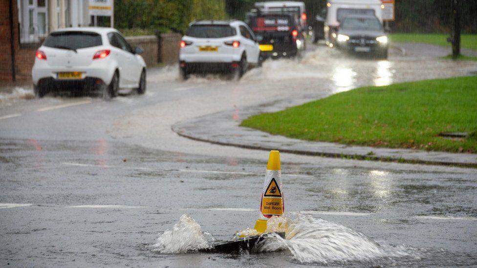 Water gushes up from a manhole in a road with a yellow cone marking cars drive through floodwater in the background