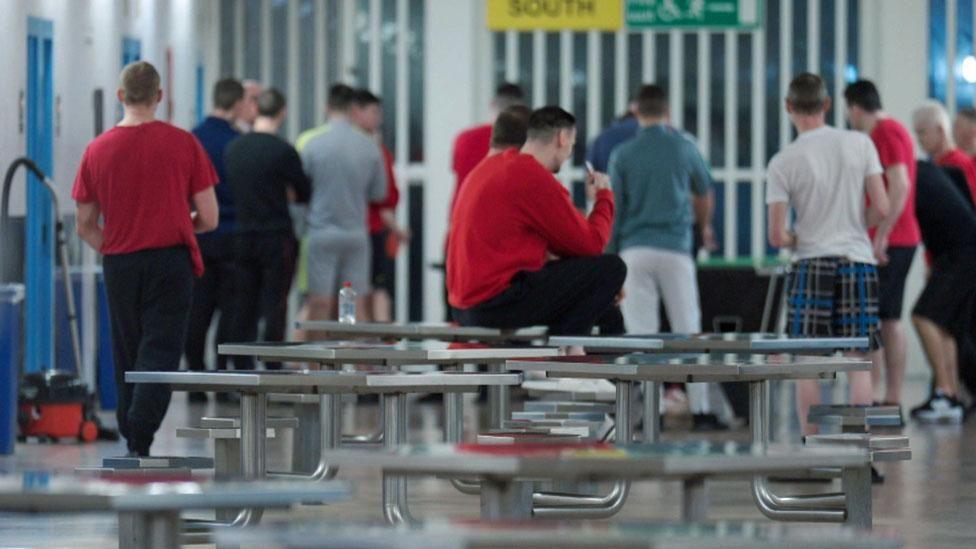 A group of prisoners in a communal area in a Scottish jail. All have their backs to the camera and one man is sitting on a table.