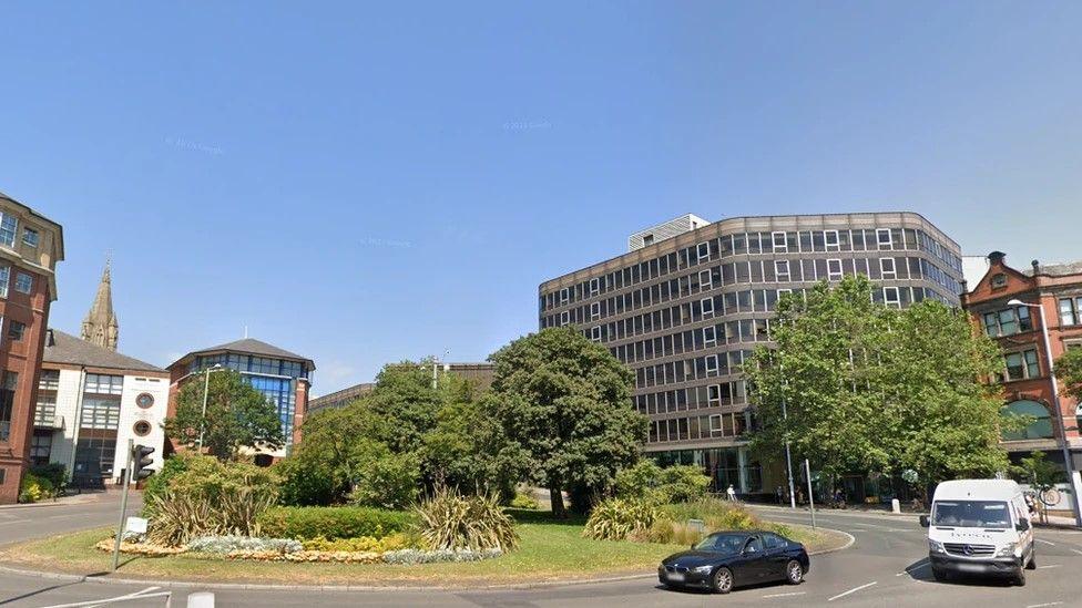 A large roundabout covered in grass and featuring trees and bushes sits in front of various buildings on Maid Marian Way