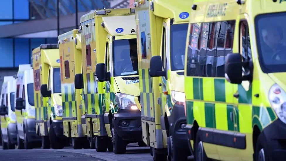 A stock photo of a line of six ambulances queue up in front of an unseen building.