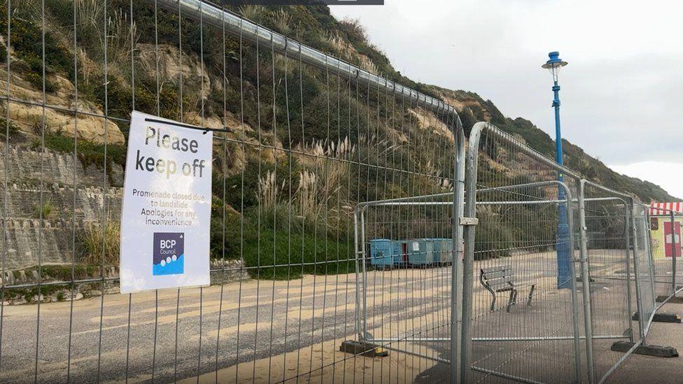 Warning sign attached to metal fencing with bench seen behind and cliffs stretching above and along the seafront.