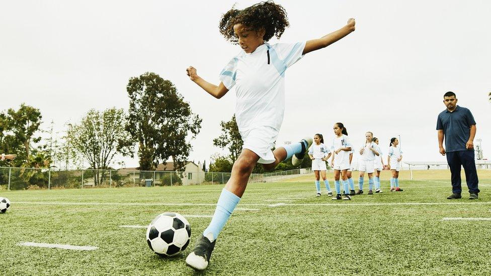 Girl kicking football with coach and rest of team in background