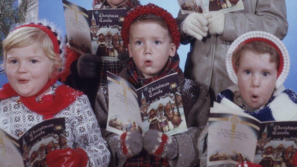 Portrait of a group of five children dressed in winter coats and hats, as they sing from a book entitled 'Christmas Carols'