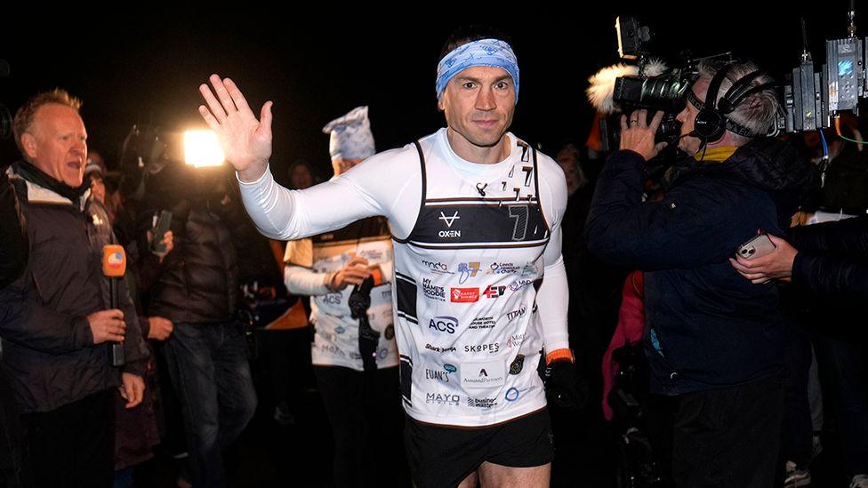 Kevin Sinfield waves as he sets off wearing a white running top and blue bandana surrounded by TV cameras