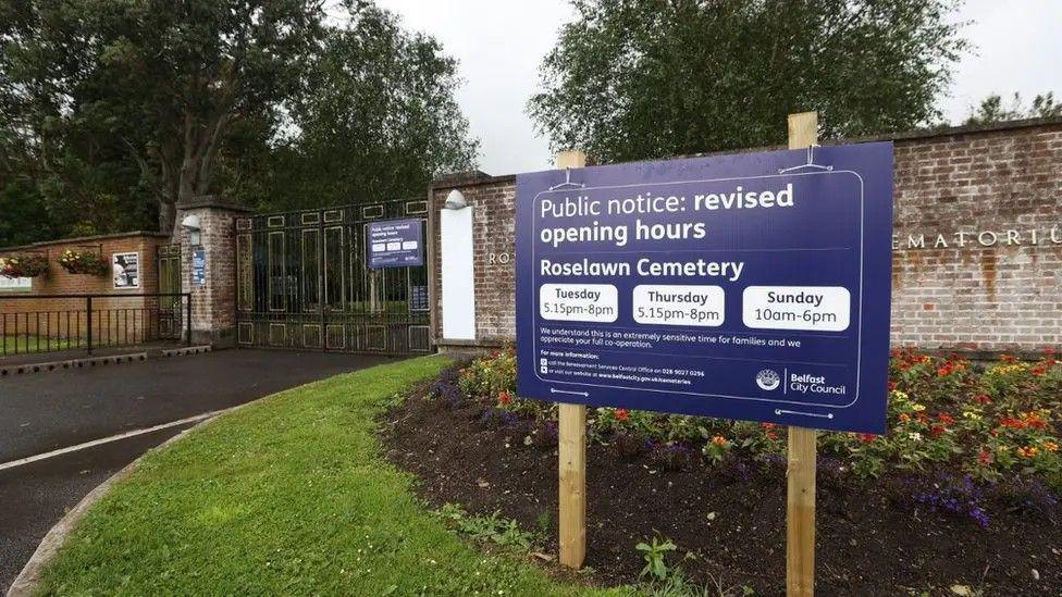 A blue sign for Roselawn Cemetery in front of black and gold gates. The opening hours are displayed. Flowers are planted beside a brick wall.