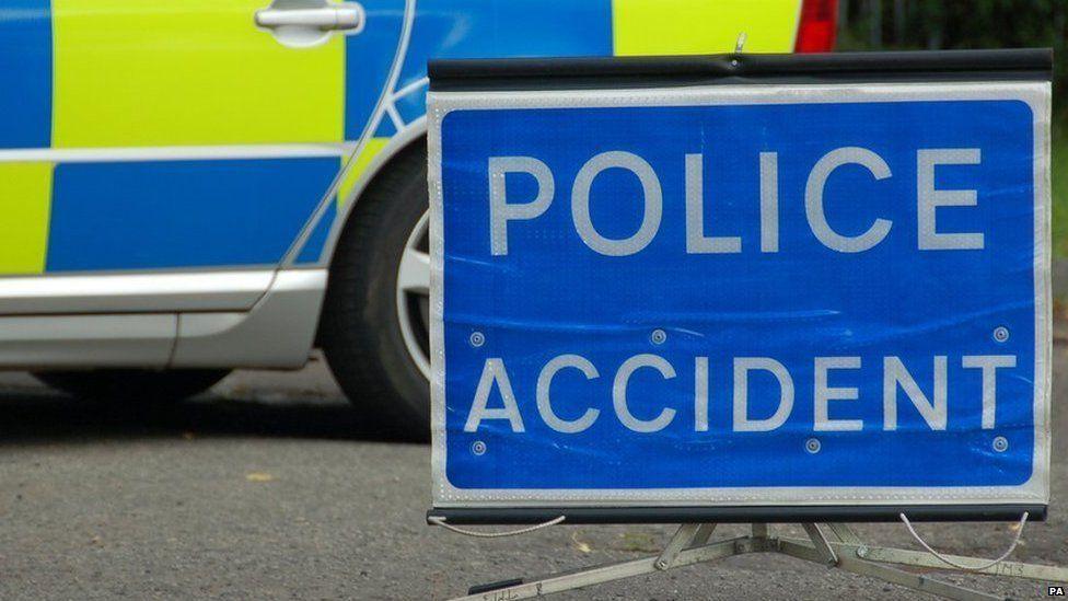 A blue and white police sign reading 'police accident'. It is on a road with a police car in the background.