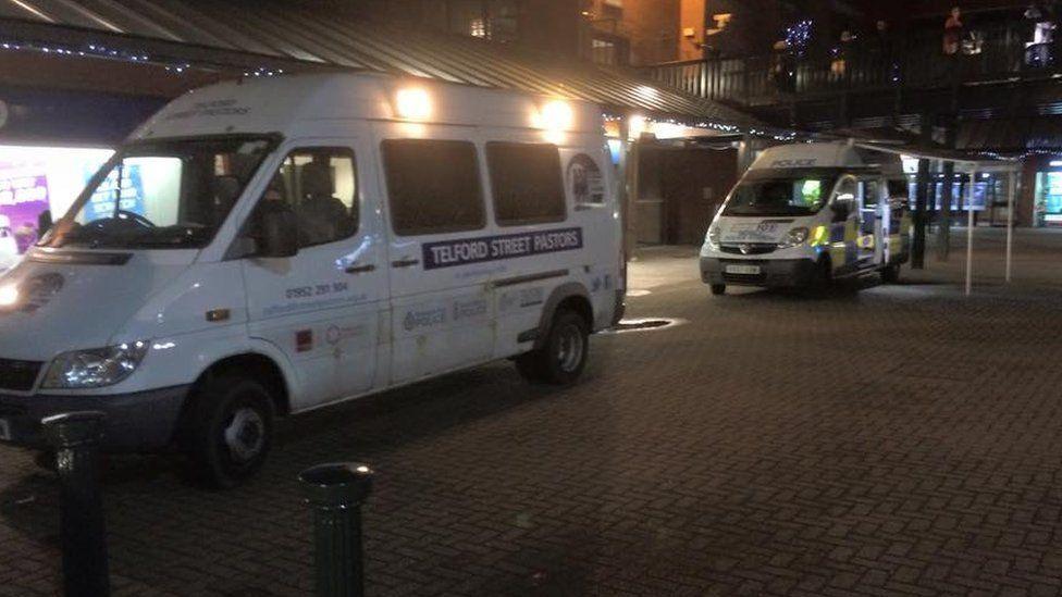 A white van that has Telford Street Pastors written on the side, parked up along a high street. Behind it a police van is also parked