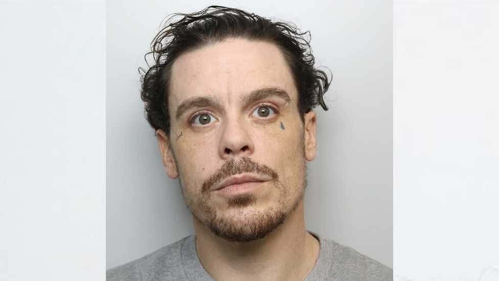 Mugshot of Luke Gleeson. He has wavy, swept back black hair and a beard. He is wearing a grey prison-issue shirt. He has small tattoos near both eyes.