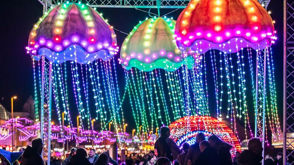 Three giant jellyfish lit up in pink, purple and green. They have long tentacles of lightss, which people can pass under.