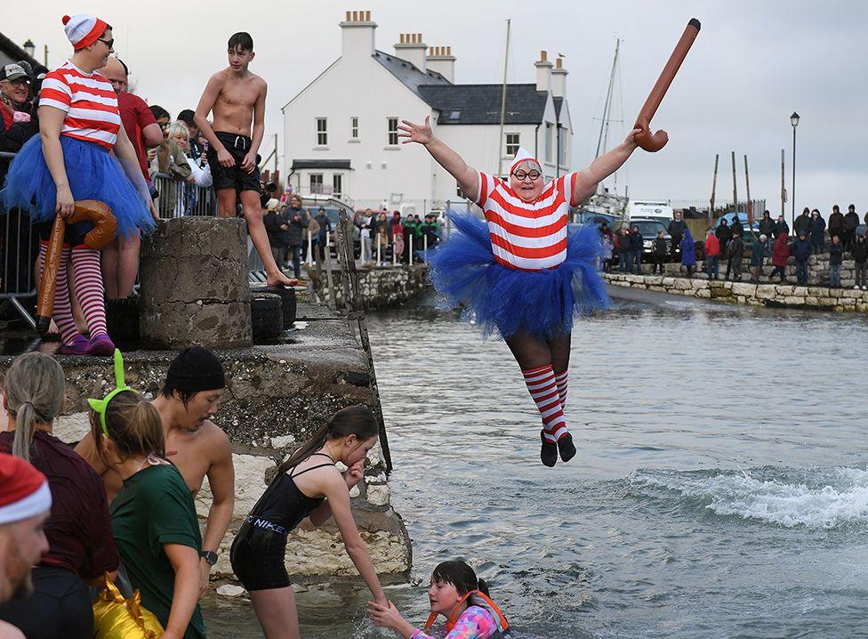 A woman, dressed in a 'Where's Wally?' costume and holding an inflatable walking stick, jumps into the water as people stand and watch on the harbourside - 1 January 2025.