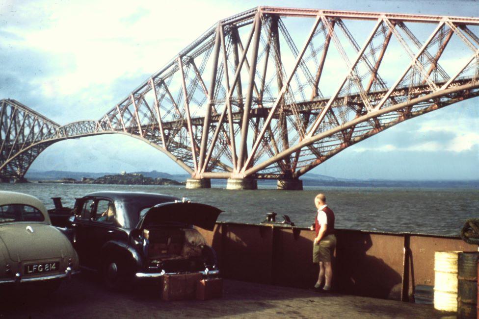 Man next to old car with its boot open with large iron bridge over water in the background