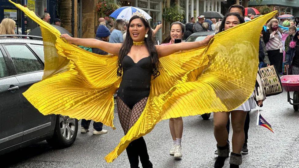 Woman in a black body and tights with bright yellow winged costume and a crown in Pride parade