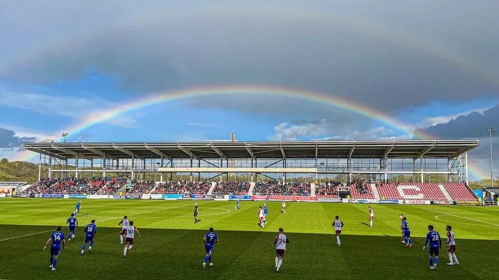 Half-built football stand on match day with a rainbow in the sky above it