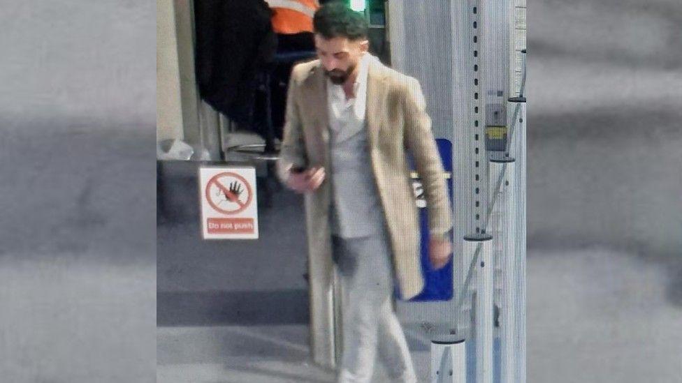 A man is wearing a white shirt and light coloured trousers. He is wearing a coat. He has black hair and facial hair and is looking at a phone he is carrying.