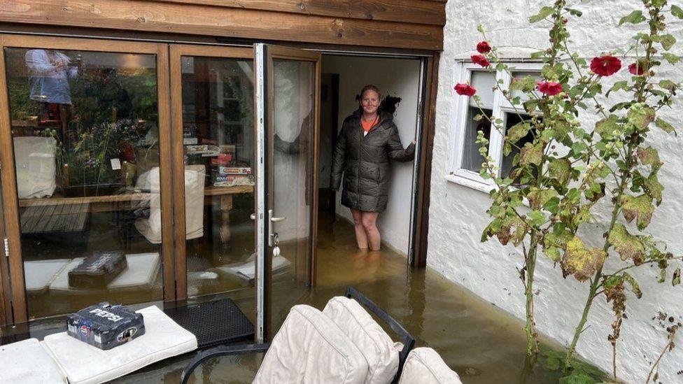 Garden furniture on a patio with brown water flooding into the doors of Nicky Vernede's home. She is standing in the water, wearing a coat, with water up to her bare shins