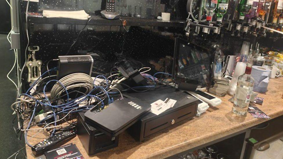 The ransacked bar at the club. There is a broken, electronic till with a mass of tangled electrical wires. Optics can be seen in the background along with opened spirit bottles on the counter.