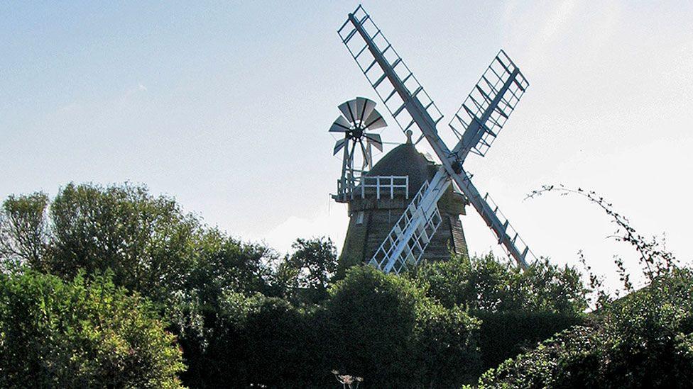 The top half of Fulbourn windmill above trees. It shows its white sails and fantail attached to the base of its conical top. Beneath it can be seen timbered walls.