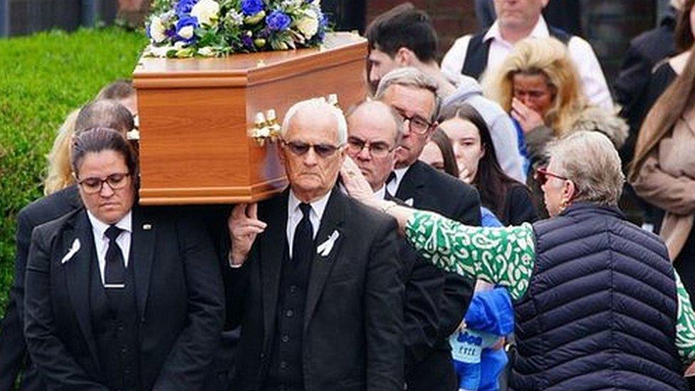 Max's coffin being carried out of Imperial Sports Ground as a crowd gathers around it to pay their respects. There is a bunch of purple and white flowers laid on top