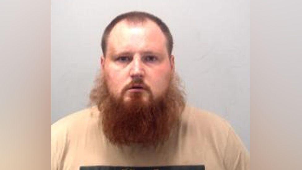 A man with a large beard and a cream T-shirt staring at the camera