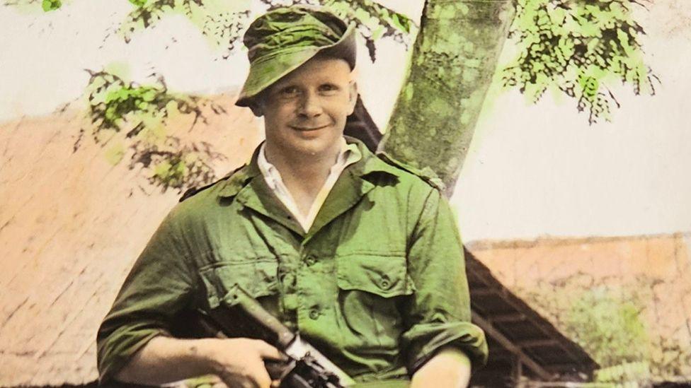 Arthur Symes standing in front of a tree in green army uniform holding a rifle