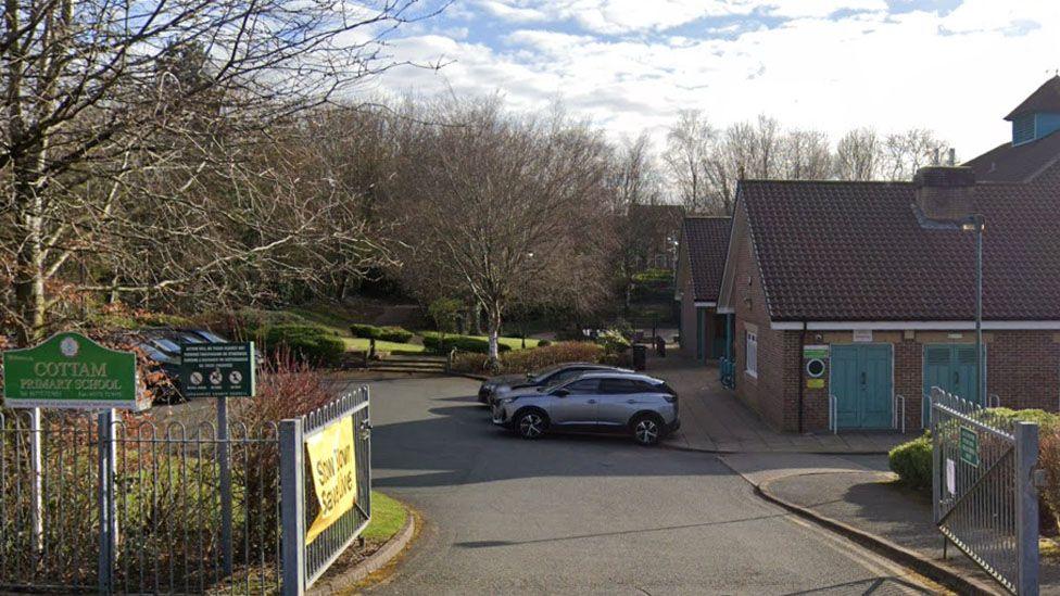 Street view of Cottam Primary School with a green sign to the left, a view of a car park and a single-storey building to the right. Trees are seen on a slope behind the school. 