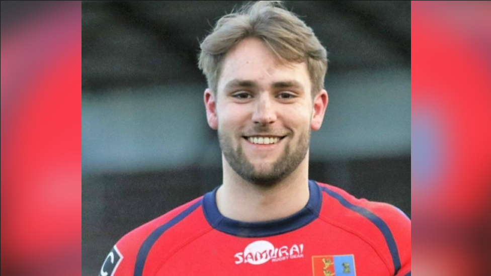 Jon Devereux is seen from his shoulders up wearing a red rugby uniform. He is smiling.