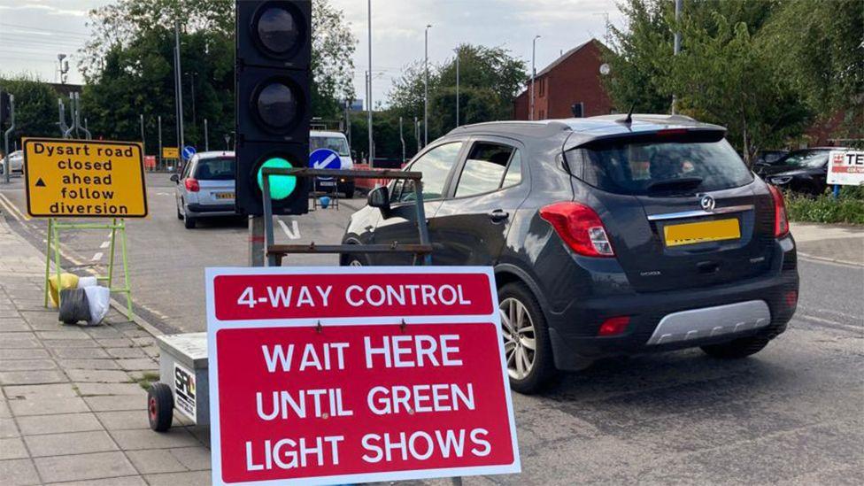 A black car passes temporary traffic lights with a diversion sign saying Dysart Road closed. In front of the temporary traffic light is a red sign saying: "4-Way control. Wait here until green light shows"