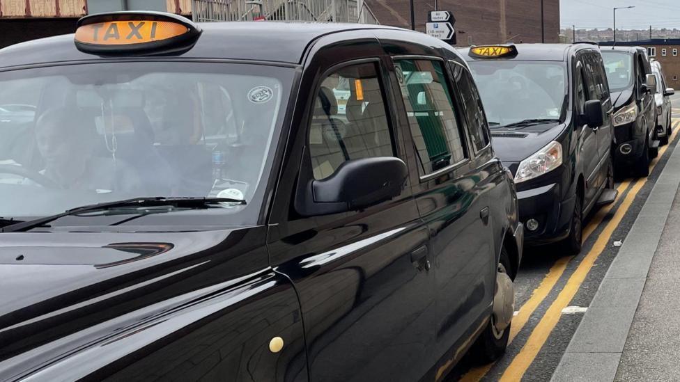 A row of black taxis with their lights on, parked on a double yellow line on the side of the road.