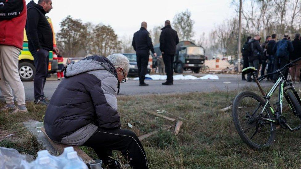 Local residents at the site of a military strike in the village of Hroza, north-eastern Ukraine on 5 October 2023. A man is seen in the foreground sitting on a concrete ledge amid a patch of grass.