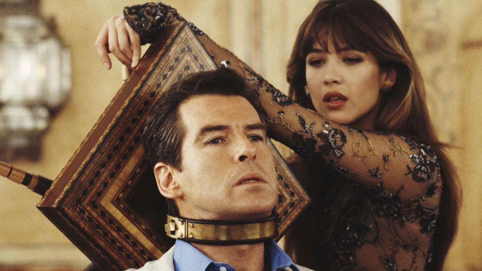 A still from The World is Not Enough. On the left is Pierce Brosnan as Bond, with short black hair, wearing a blue shirt under a cream jacket. Around his neck is a brass coloured band, imprisoning him on the headrest of a chair. Resting her arm on the headrest is Sophie Marceau as Elektra King. She has long dark hair and is wearing a net dress covered in embroidery.