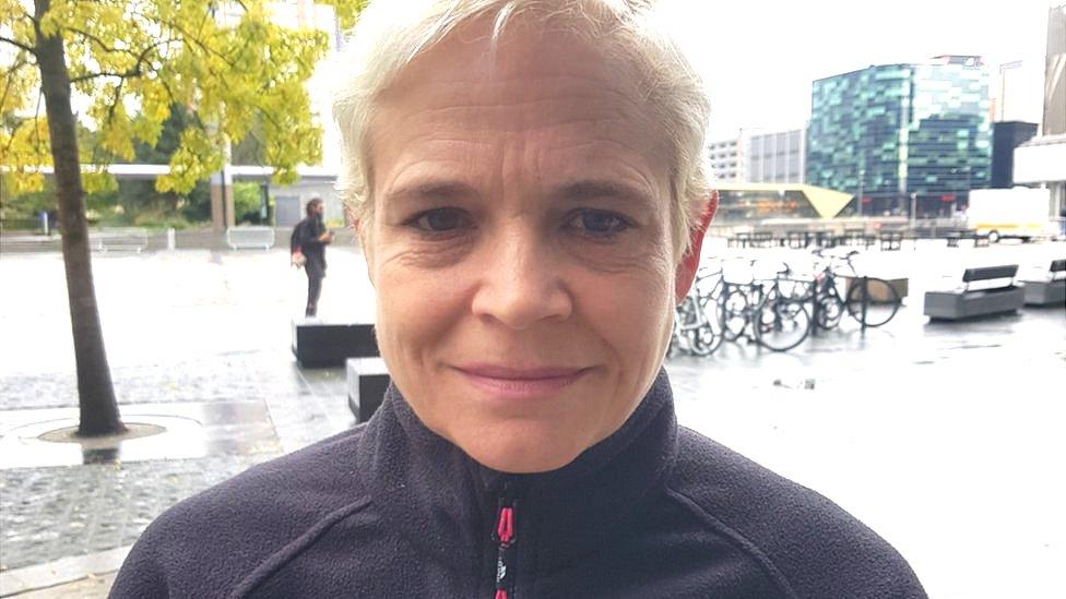 A head and shoulders shot of a woman with short, dyed white hair and a black fleece top standing outside in front of modern glass buildings and a bicycle rack.