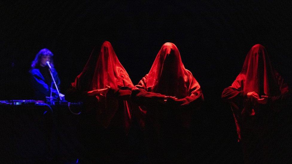 Three women draped in red veils, with a musician in the background