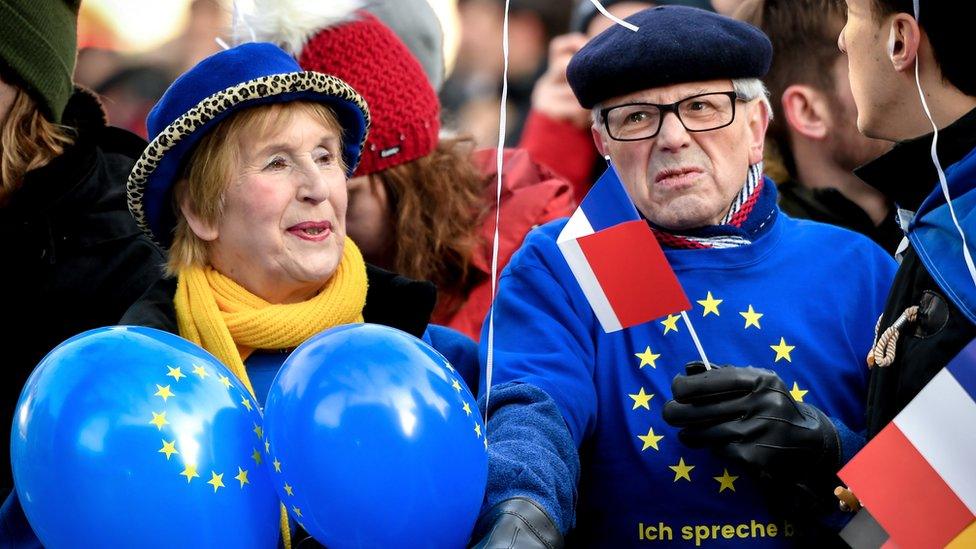 A man and woman dressed int eh bright blue and yellow colours of the EU stand at a barrier, holding balloons and the French flag on a stick