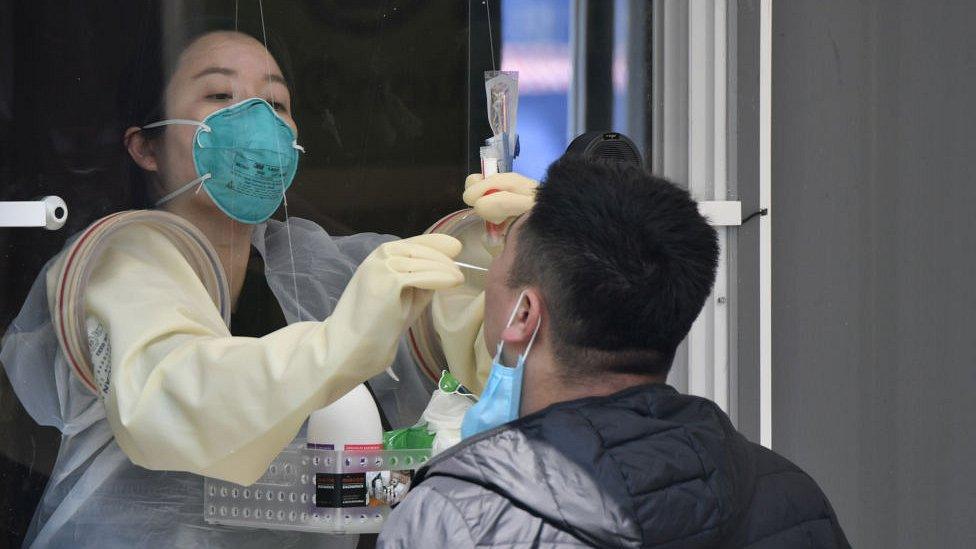 A woman wearing a mask doing a throat swab on a man through a window