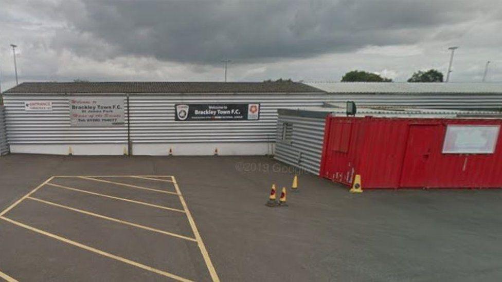 Single-storey corrugated iron buildings, mostly grey but with one red painted wall to the right.  A sign says "Brackley Town FC".