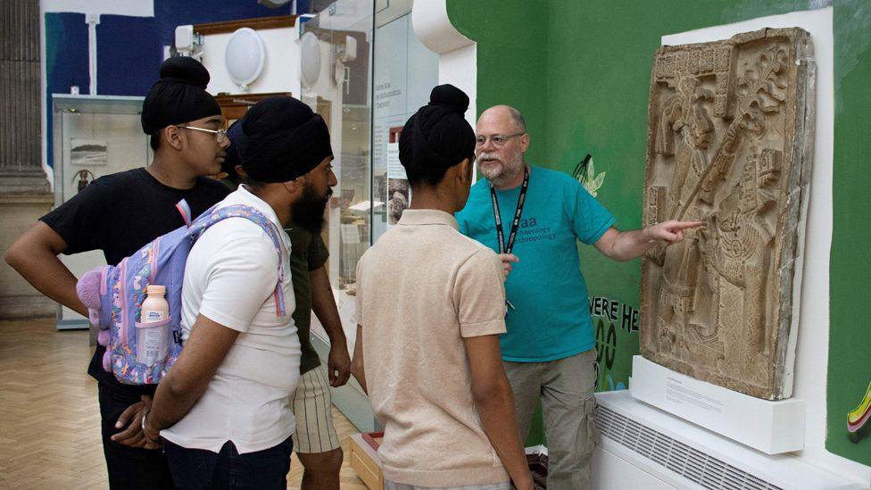 Several people looking at an object at the Museum of Archaeology and Anthropology, in Cambridge. One man has a lanyard on pointing at the item, and four other people are looking at it.