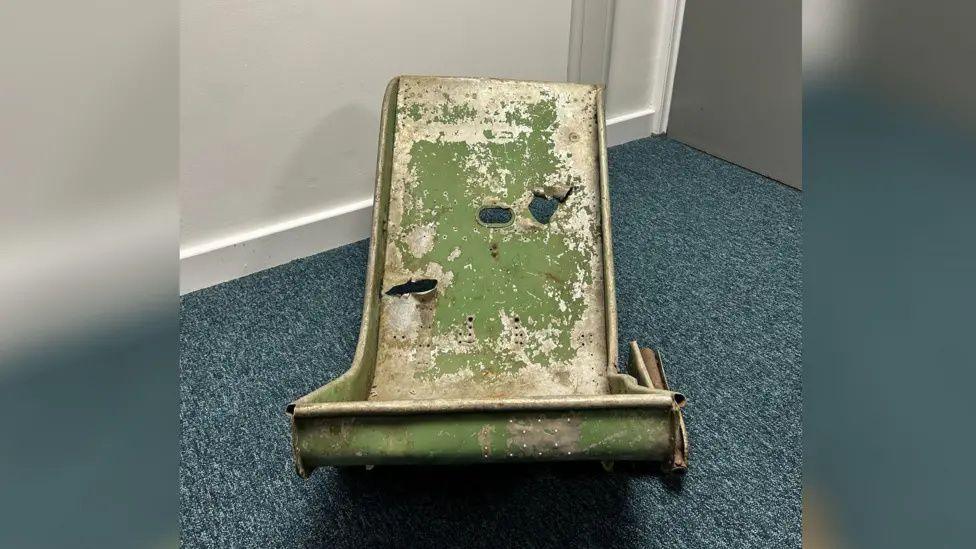 A world war 2 plane seat lies on a carpet in a simple room. Its colour is partly green but it has clearly faded into white in places, and several holes caused by shrapnell or bullets are visible