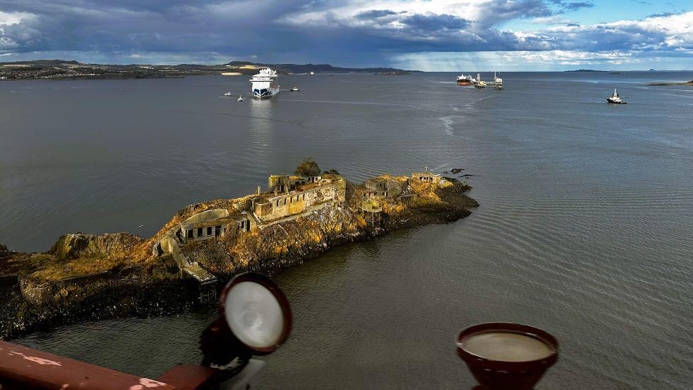 An island in the estuary which has derelict buildings on it. There are boats on the water in the background.