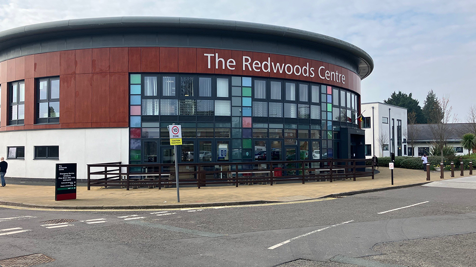 The Redwoods Centre. Built in the round, the upper part of the building is covered in red wooden panels, with the lower part in white render. The front of the building, under a large sign reading "The Redwoods Centre" features lots of glass windows