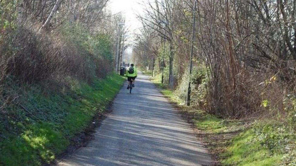 A view of the cycle path with grassy banks either side of it and a cyclist wearing a hi-vis jacket riding along it